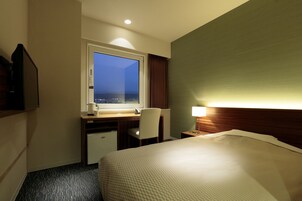 Quarto de hotel com uma cama, uma escrivaninha com cadeira, um frigorífico pequeno, uma janela com vista e uma mesa de cabeceira com abajur.