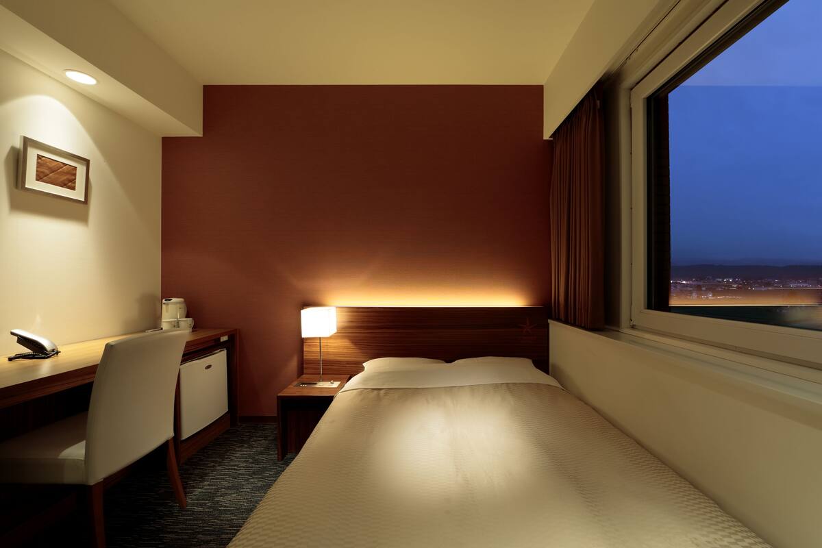 A hotel room with a bed, desk, chair, nightstand, lamp, and a window with a city view.