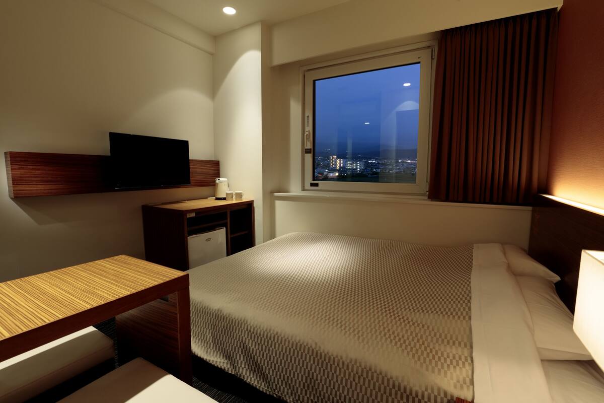 A hotel room with a bed, a desk, a television, and a window with a city view.