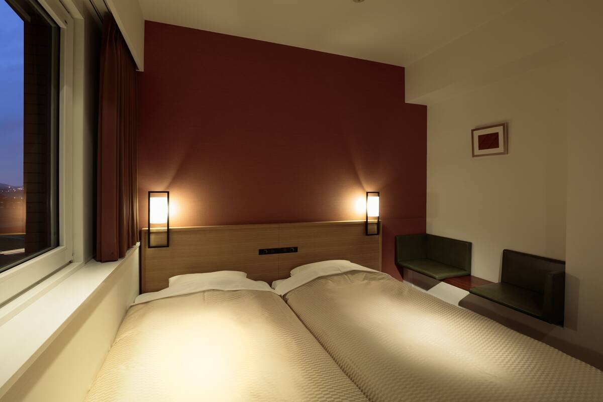 A hotel room with a bed, two bedside lamps, a window, a green chair, and a small table.