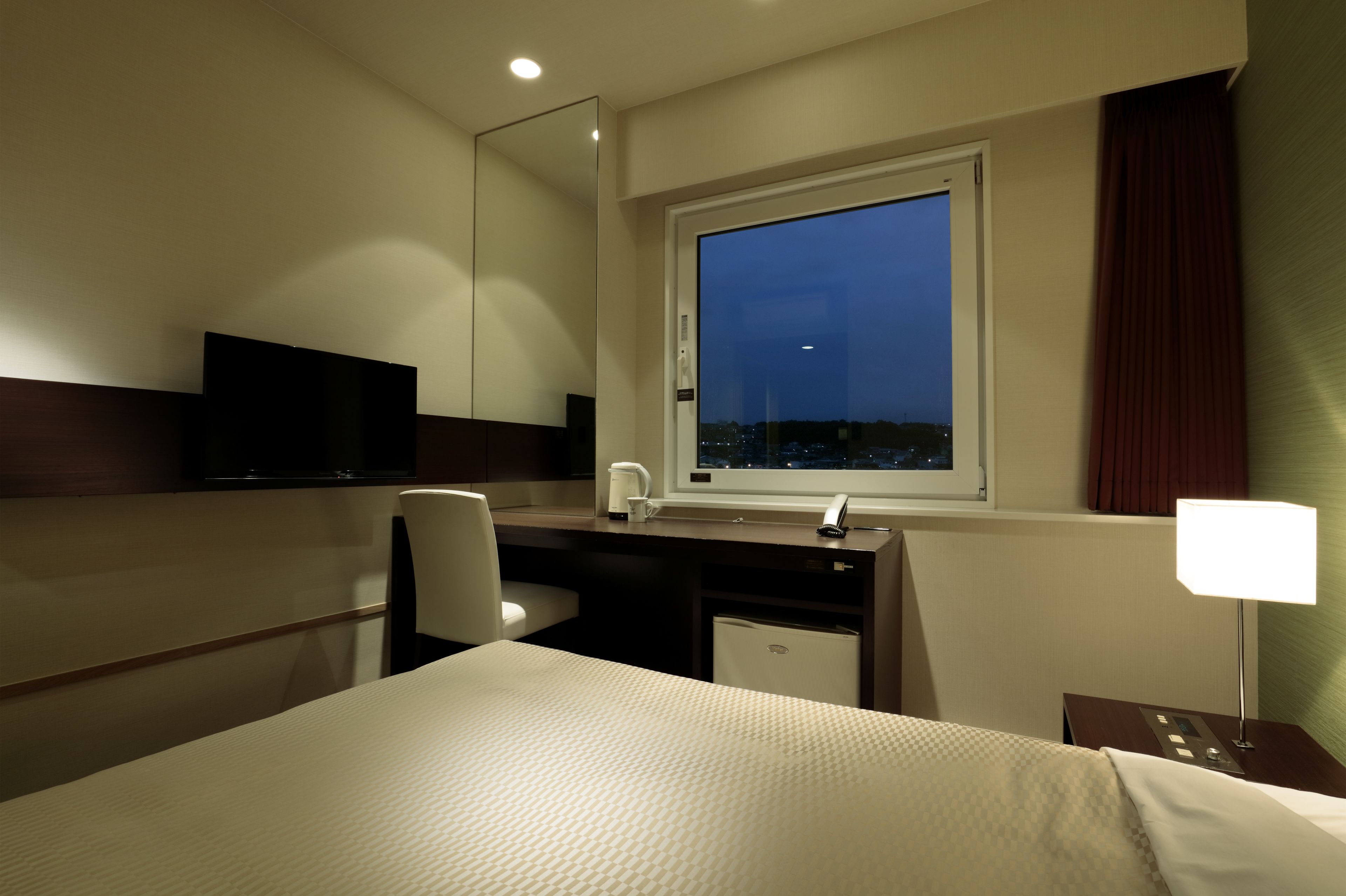 A hotel room with a bed, a desk with a chair, a television, a window with a view of the city at night, and a nightstand with a lamp.