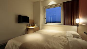A hotel room with a bed, a bedside table with a lamp, a flat-screen TV mounted on the wall, and a window with curtains.