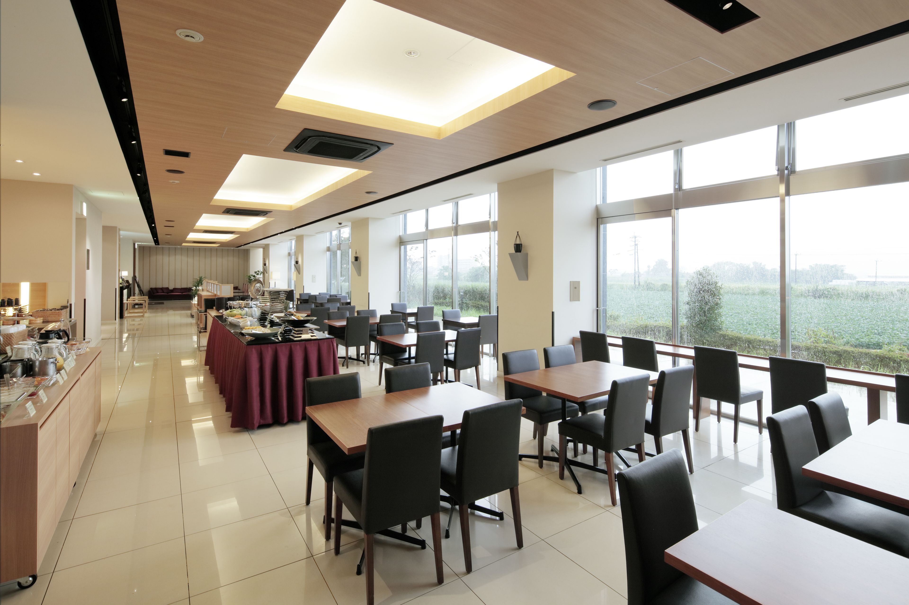 A spacious dining area with multiple tables and chairs, large windows offering a view of the outdoors, and a buffet setup on the left.