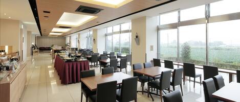 A spacious dining area with multiple tables and chairs, large windows offering a view of the outdoors, and a buffet setup on the left.
