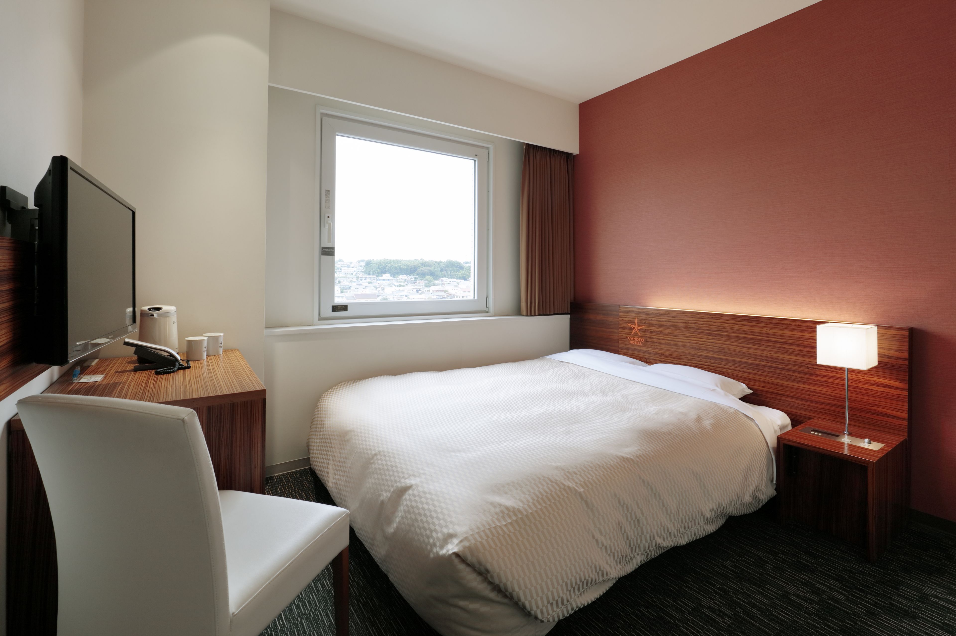 Et hotelværelse med seng, skrivebord med stol, fjernsyn, et vindue og et natbord med lampe.