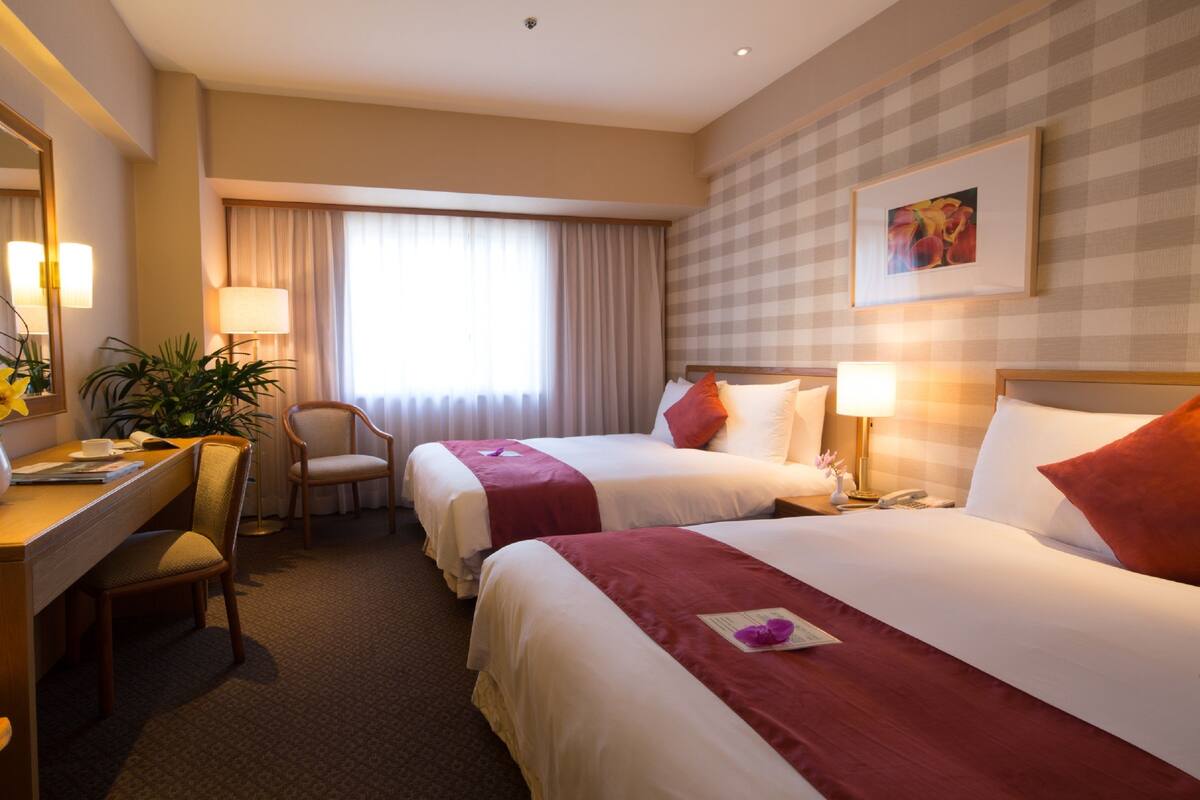 A hotel room with two beds, a desk, a chair, a lamp, and a window with curtains.