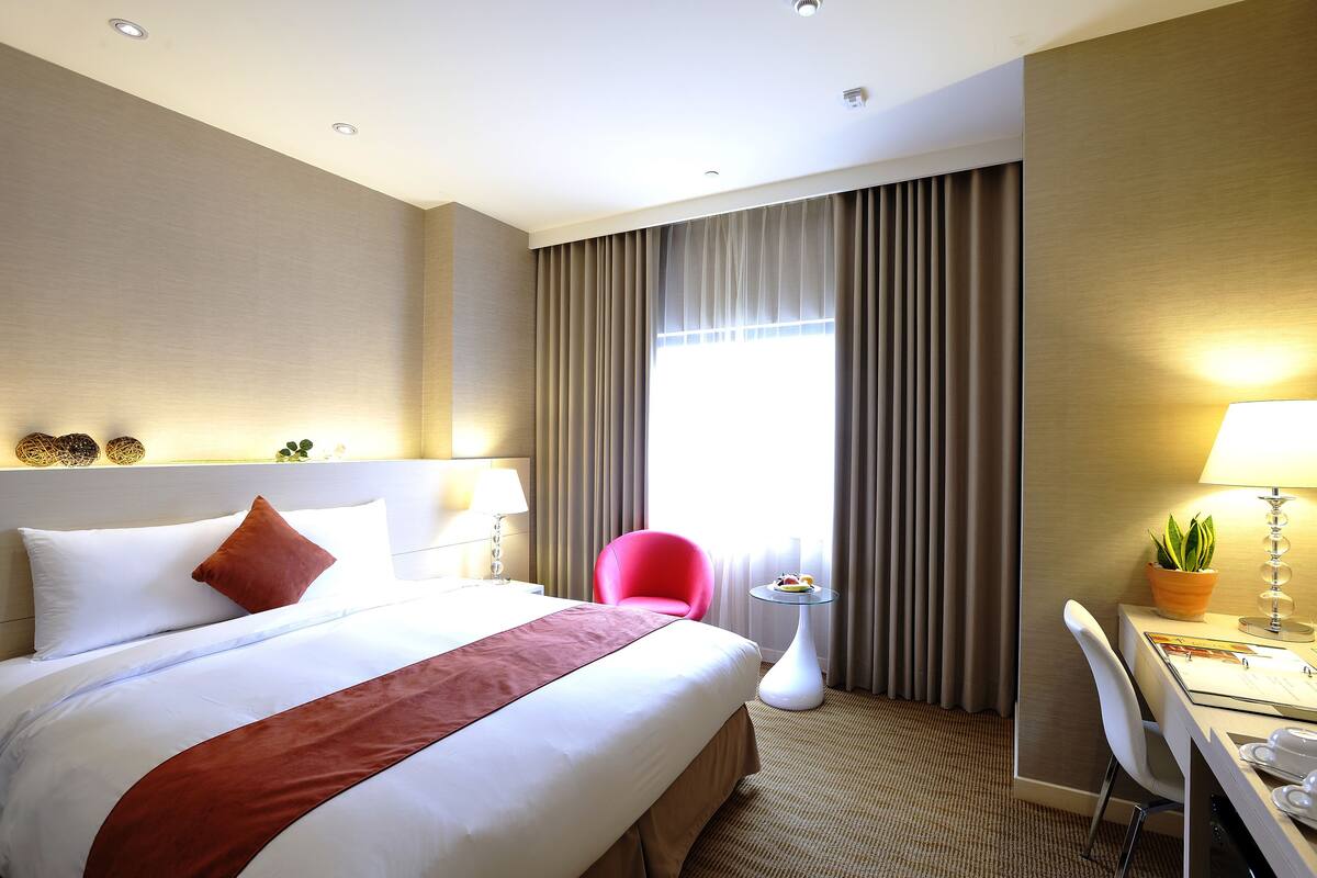 A hotel room with a large bed, a desk with a chair, a small table with a plant, and a window with curtains.