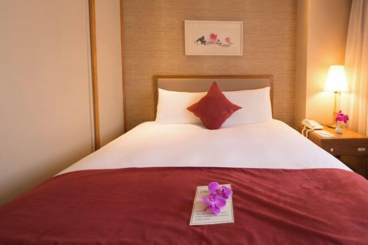 Una habitación de hotel con una cama bien hecha, un cabecero de madera, un cuadro en la pared, una mesita de noche con una lámpara y una colcha roja.