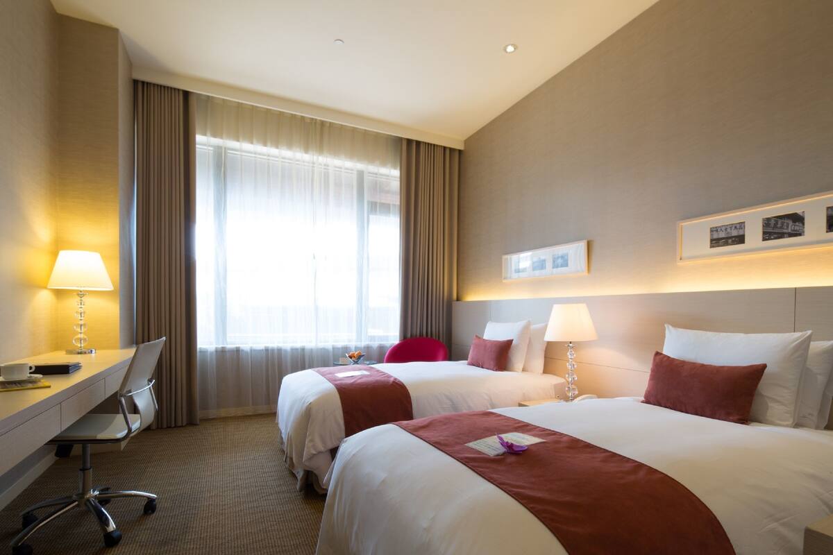A hotel room with two beds, a desk, a chair, and a large window with curtains.