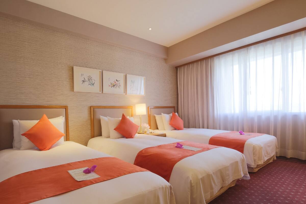 A hotel room with three beds, each with white bedding and orange pillows. There are framed artworks on the wall, a large window with sheer curtains, and a red carpeted floor.