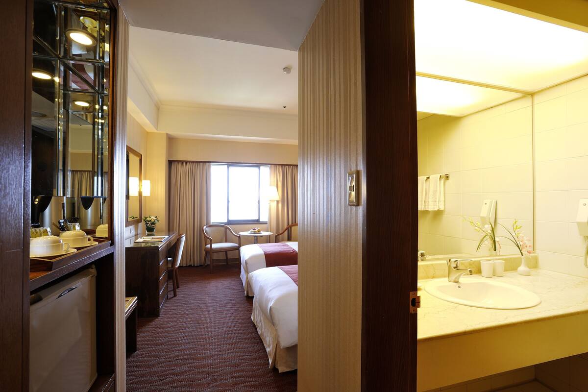 A hotel room with a desk, two beds, a bathroom with a sink and mirror, and a window with curtains.