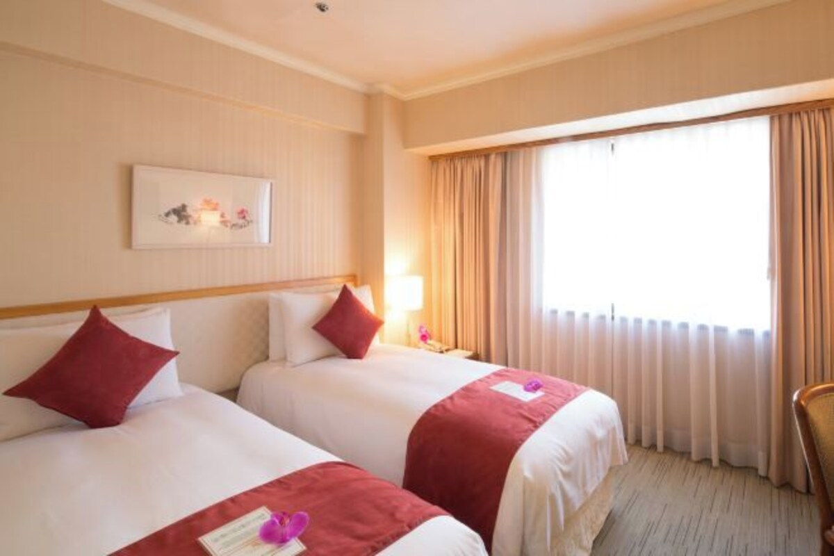 Habitación de hotel con dos camas, un ventanal con cortinas, una mesita de noche con lámpara y un cuadro en la pared.