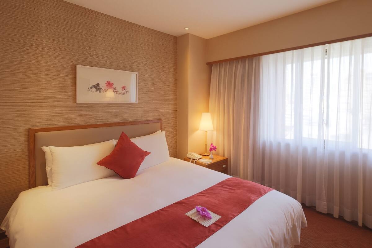 A hotel room with a large bed, a bedside table with a lamp, a telephone, and a window with sheer curtains.