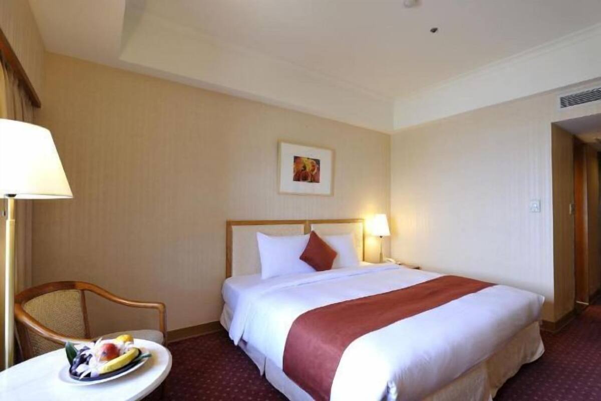 A hotel room with a neatly made bed, a round table with a fruit plate, and a lamp.
