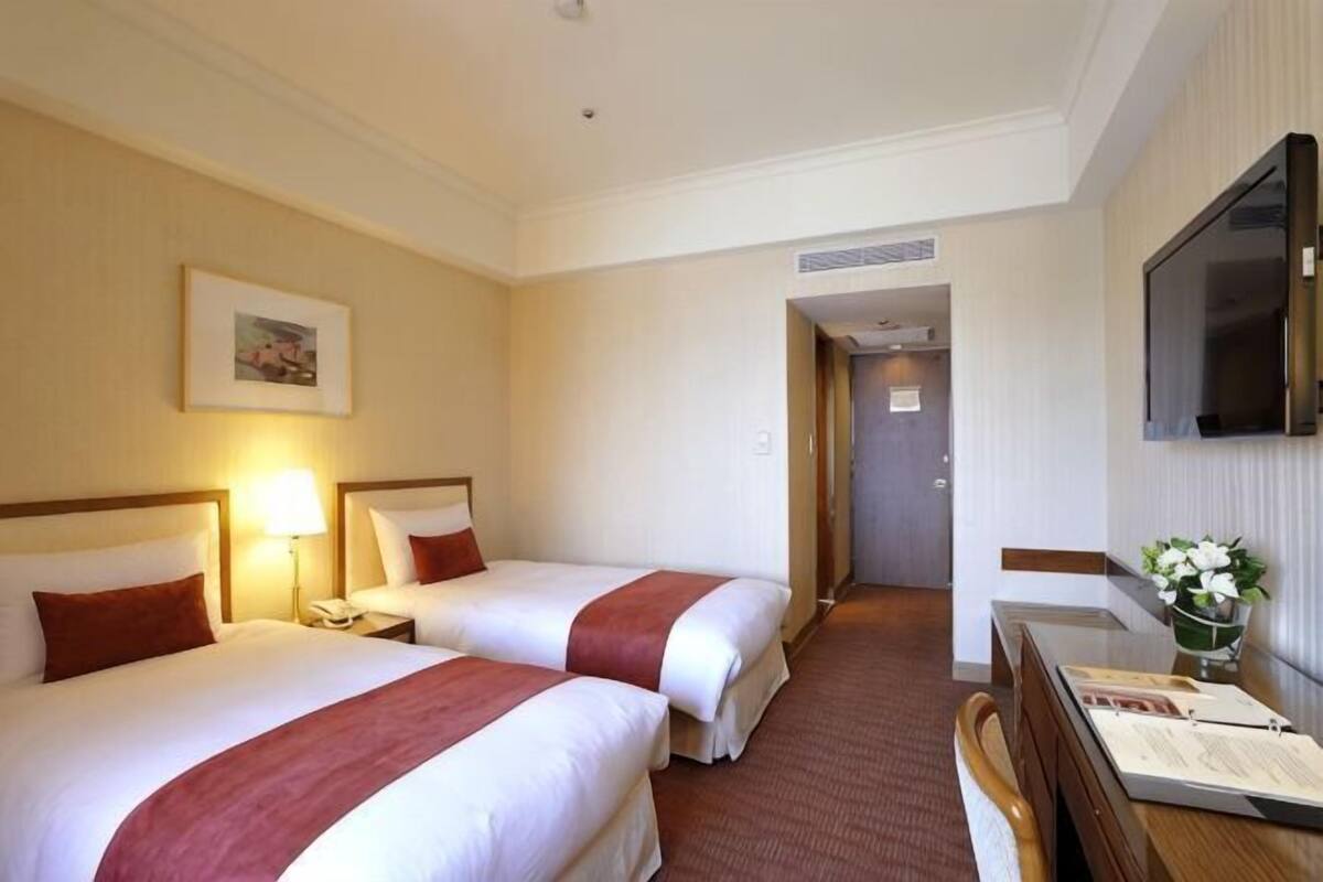 A hotel room with two beds, a desk, a chair, a TV, and a door leading to another room.