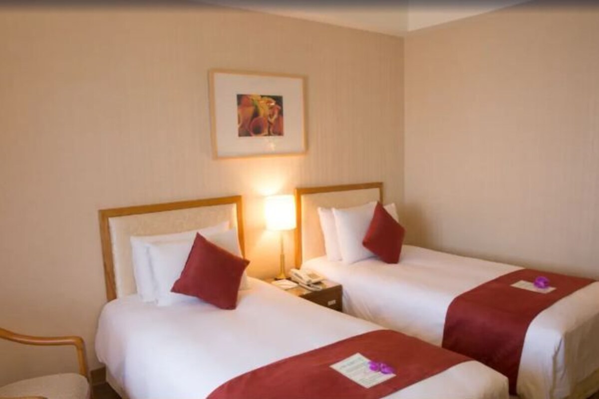 Una habitación de hotel con dos camas, cada una con ropa de cama blanca y detalles rojos, una mesita de noche con lámpara, y un cuadro en la pared.