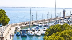 Un puerto deportivo con barcos amarrados a un muelle de piedra, un faro al fondo y una vista despejada del agua.
