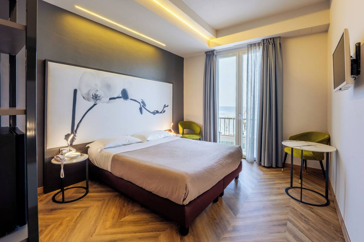 A modern hotel room with a large bed, a bedside table, a chair, a small table, and a large window with curtains.