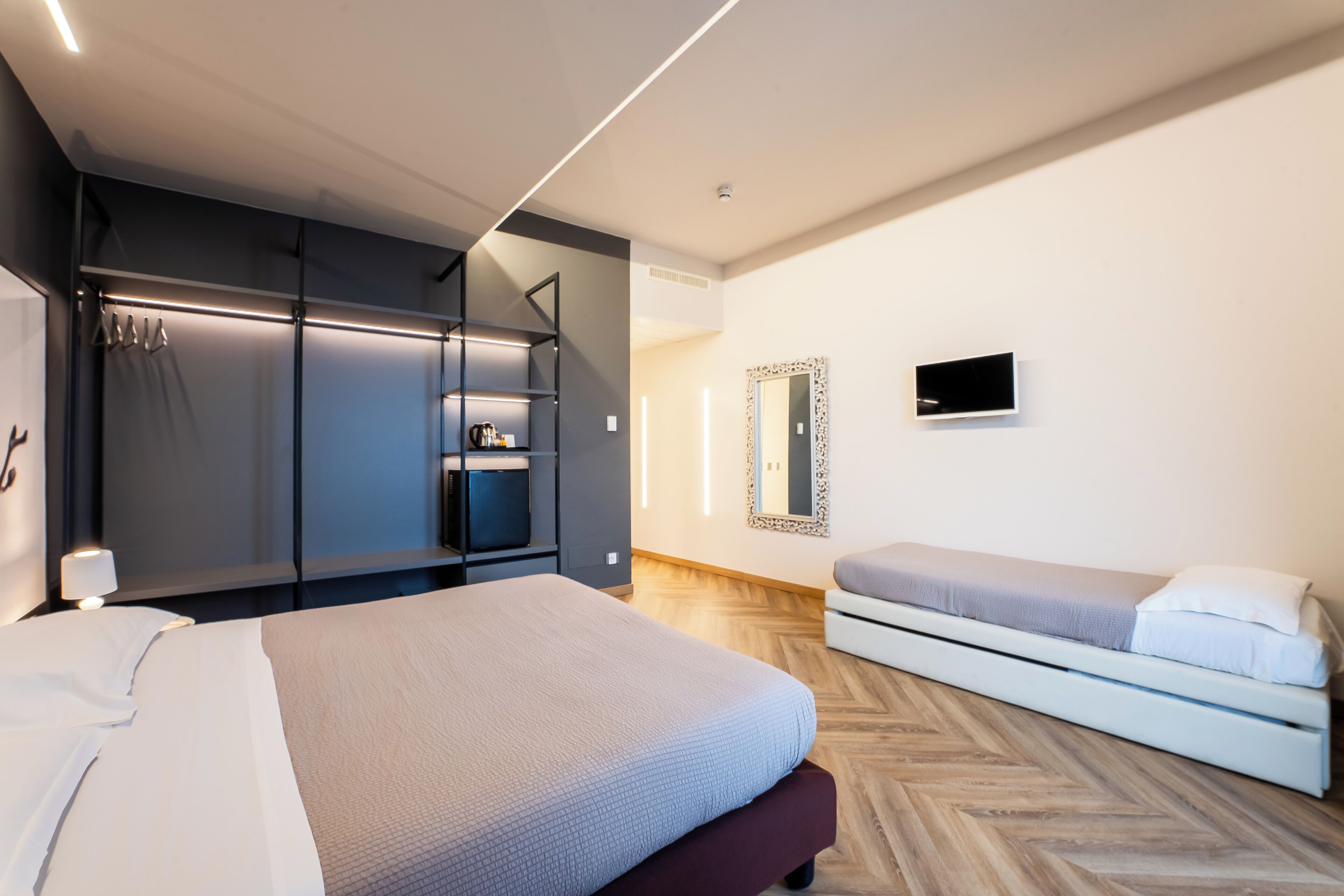 A modern hotel room with a large bed, a smaller bed, a wardrobe, a mirror, and a flat-screen TV.