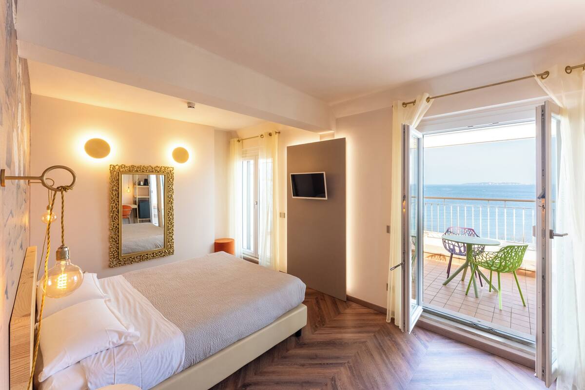 A bedroom with a bed, bedside lamps, a TV, and a balcony with chairs.