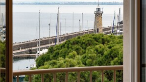 Un balcón con vista a un faro, un muelle con barcos y un área verde con árboles.