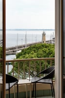 A balcony with a view of a lighthouse, a pier with boats, and a green area with trees.