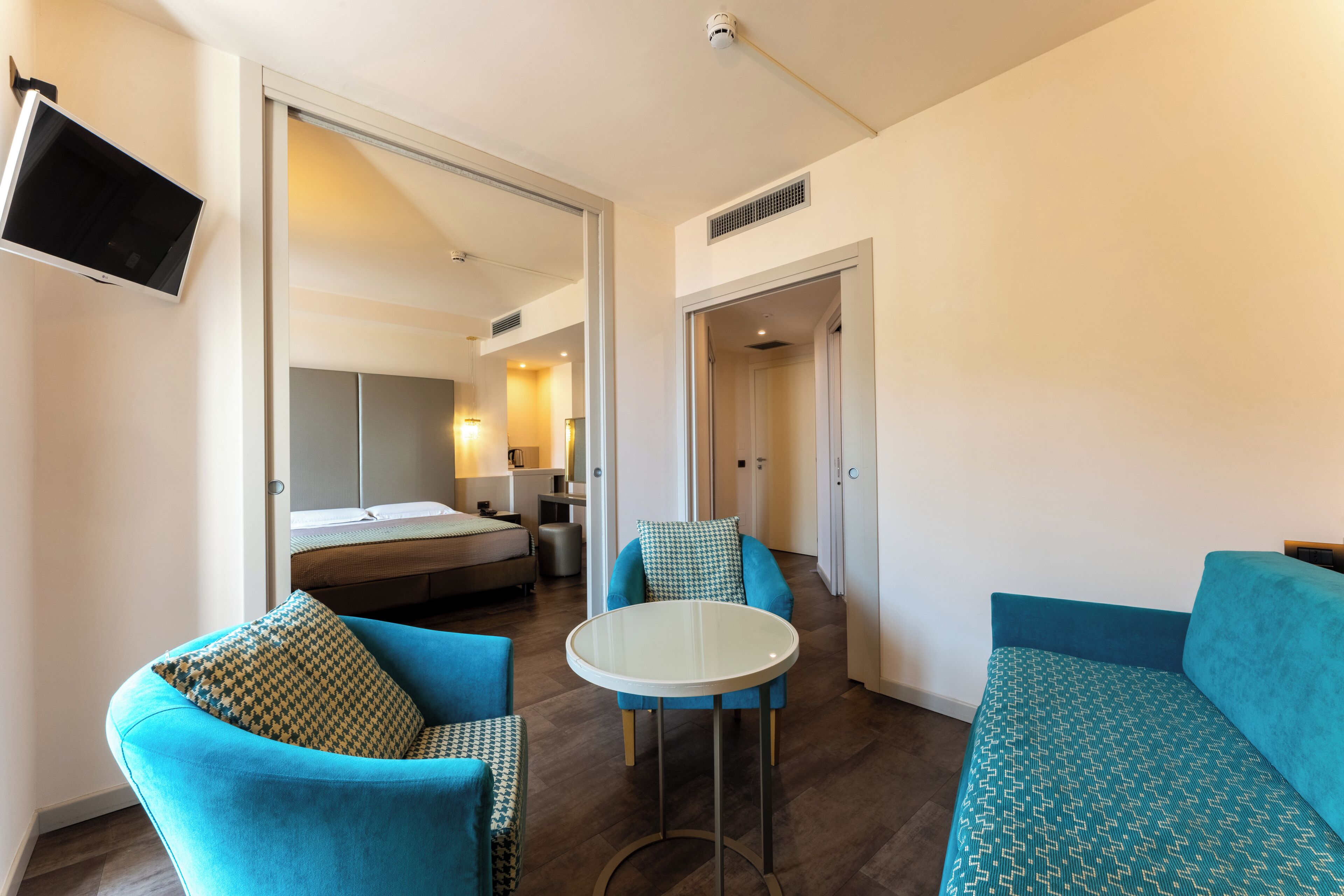 Ein modernes Hotelzimmer mit einem Bett, einem runden Tisch, zwei blauen Sofas und einem an der Wand montierten Fernseher.