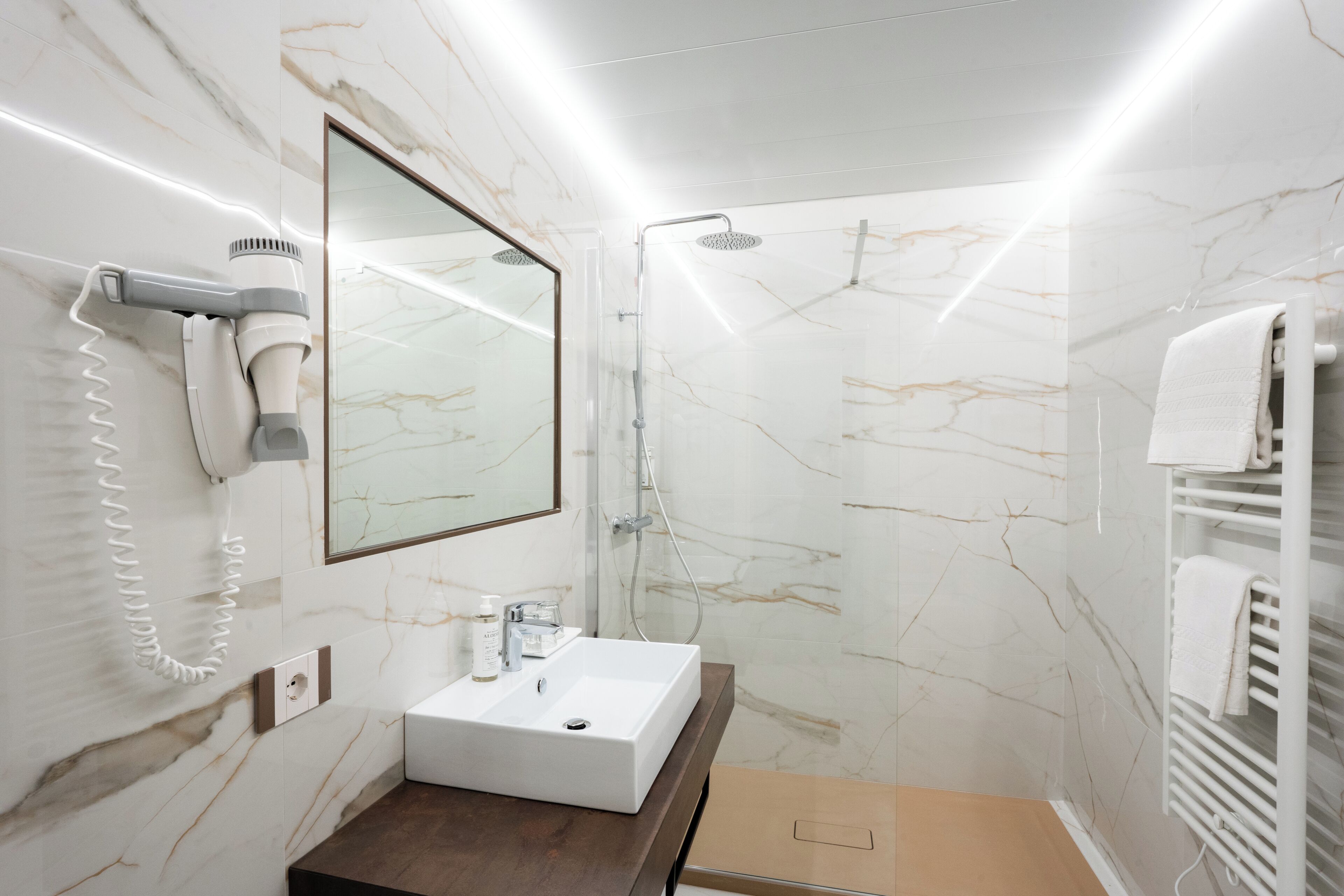 A bathroom with a marble wall, a sink, a mirror, a hairdryer, and a shower.