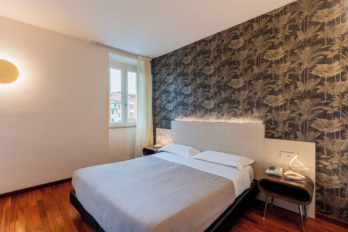 A neatly made bed with white linens, a bedside table with a lamp, and a wall with a leaf-pattern wallpaper.