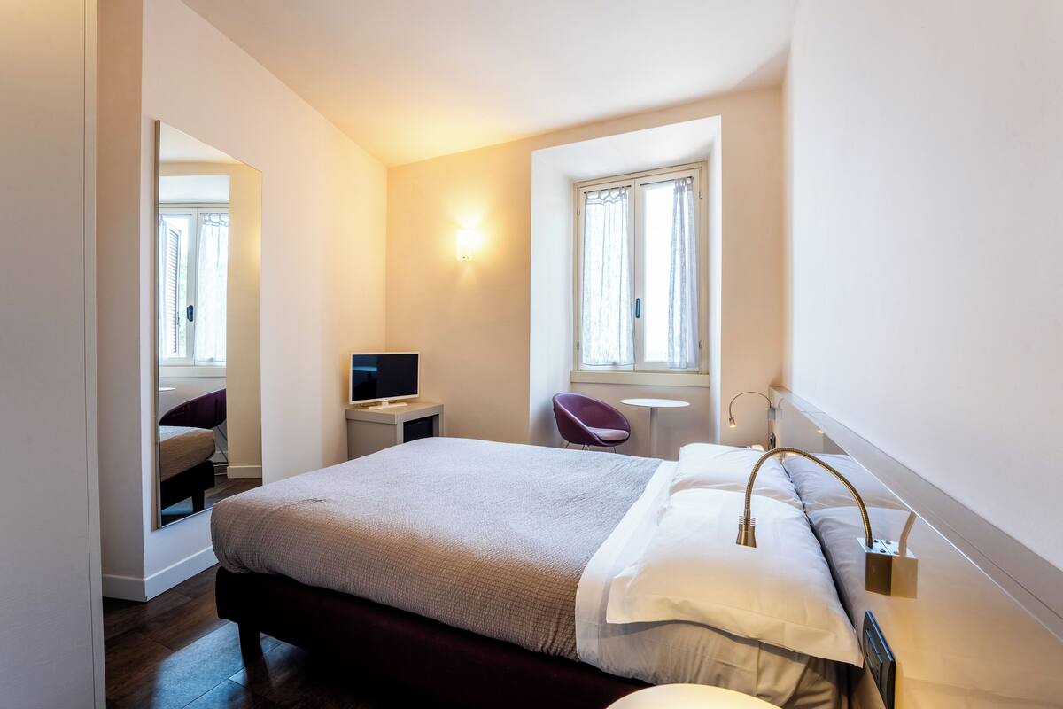 A hotel room with a bed, a desk with a computer, a chair, and a window with curtains.