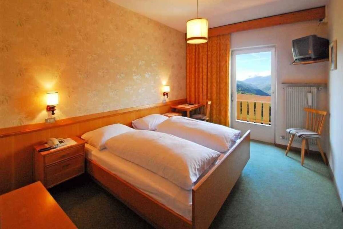 A hotel room with a double bed, bedside lamps, a wooden headboard, a small table, a chair, a TV mounted on the wall, and a view of mountains through the window.