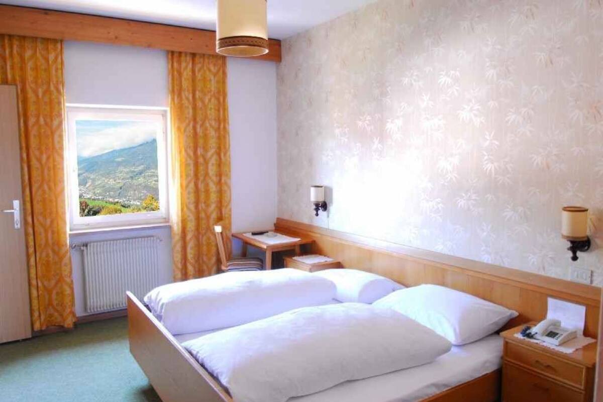 A double bed with white linens, a bedside table with a lamp, a nightstand with a phone, and a view of a mountainous landscape through a window with yellow curtains.