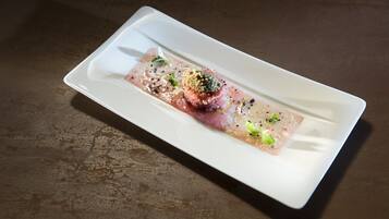 Un plato rectangular blanco con un guiso de pescado crudo, adornado con hierbas verdes y semillas negras, sobre una superficie marrón texturizada.