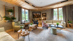 Una sala de estar acogedora con techo de madera, ventanas amplias, un sofá, sillones, una mesita de centro y plantas en macetas.