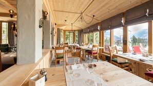 Un interior de madera con mesas y sillas, amplios ventanales con vistas a la montaña, y lámparas colgantes.
