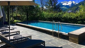 Una piscina con sillones de descanso y una pérgola, rodeada de vegetación y montañas al fondo.