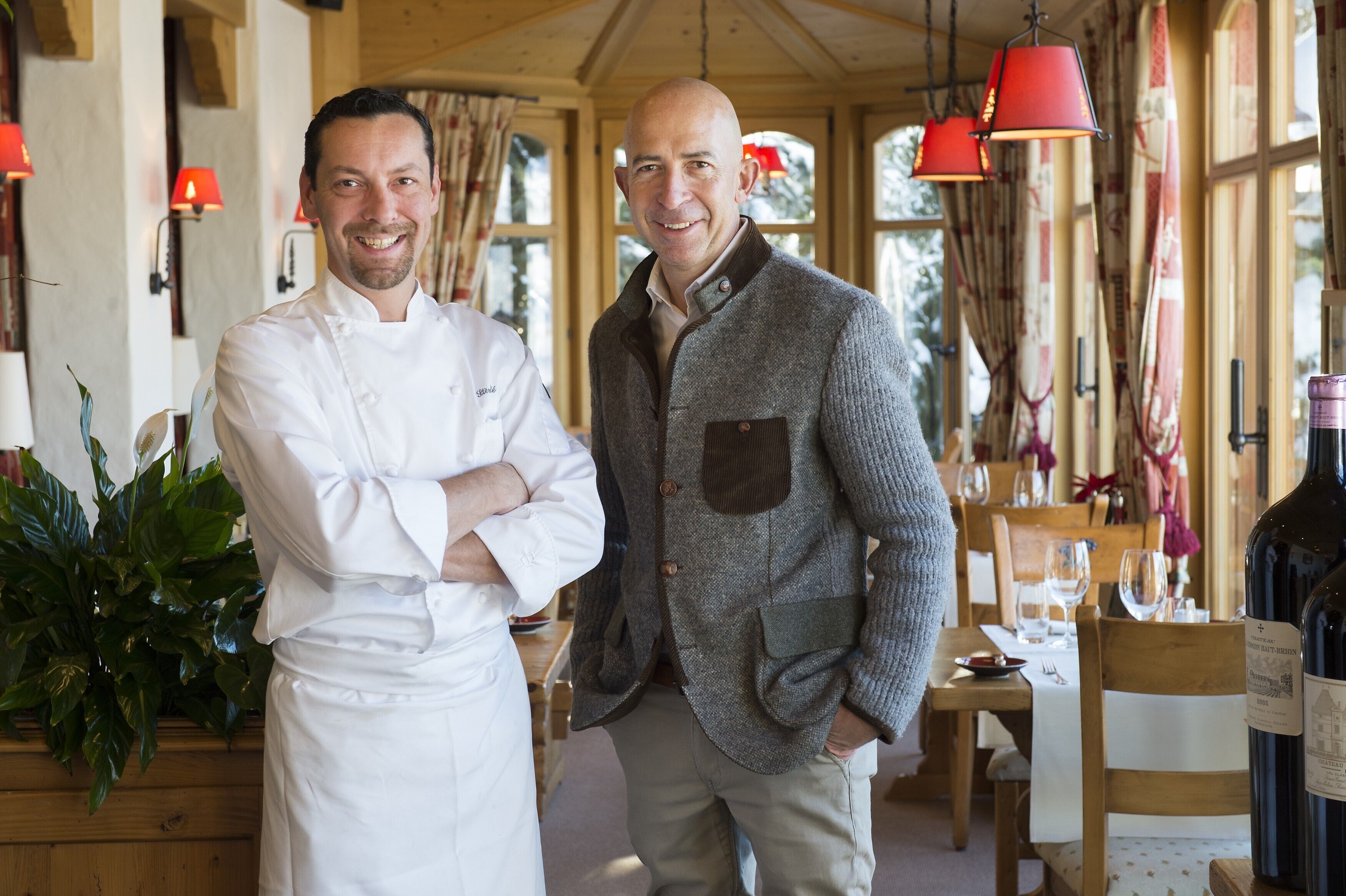 Two men in a restaurant with wooden interiors, red hanging lamps, and a table set with wine glasses.
