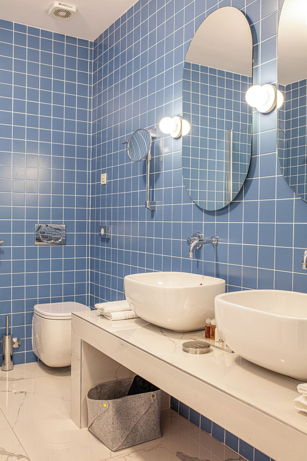 Un baño con paredes de azulejos azules, dos lavamanos, un inodoro y un espejo.
