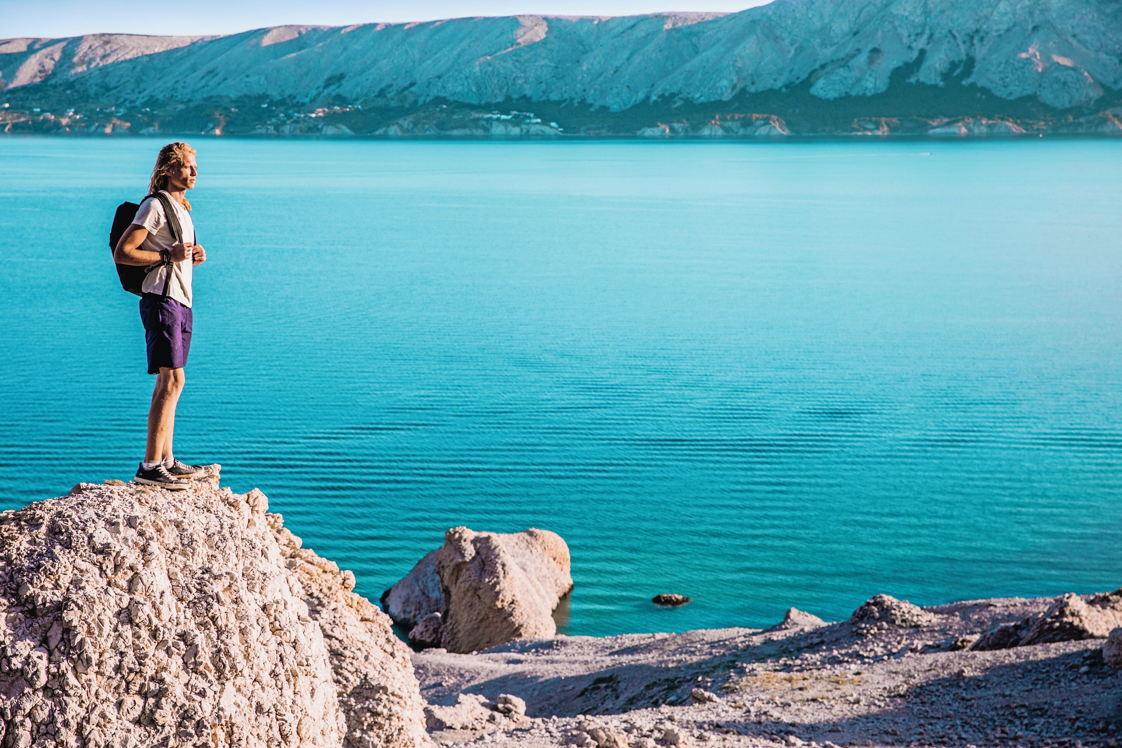 A person standing on a rocky outcrop by a calm body of water with mountains in the background.