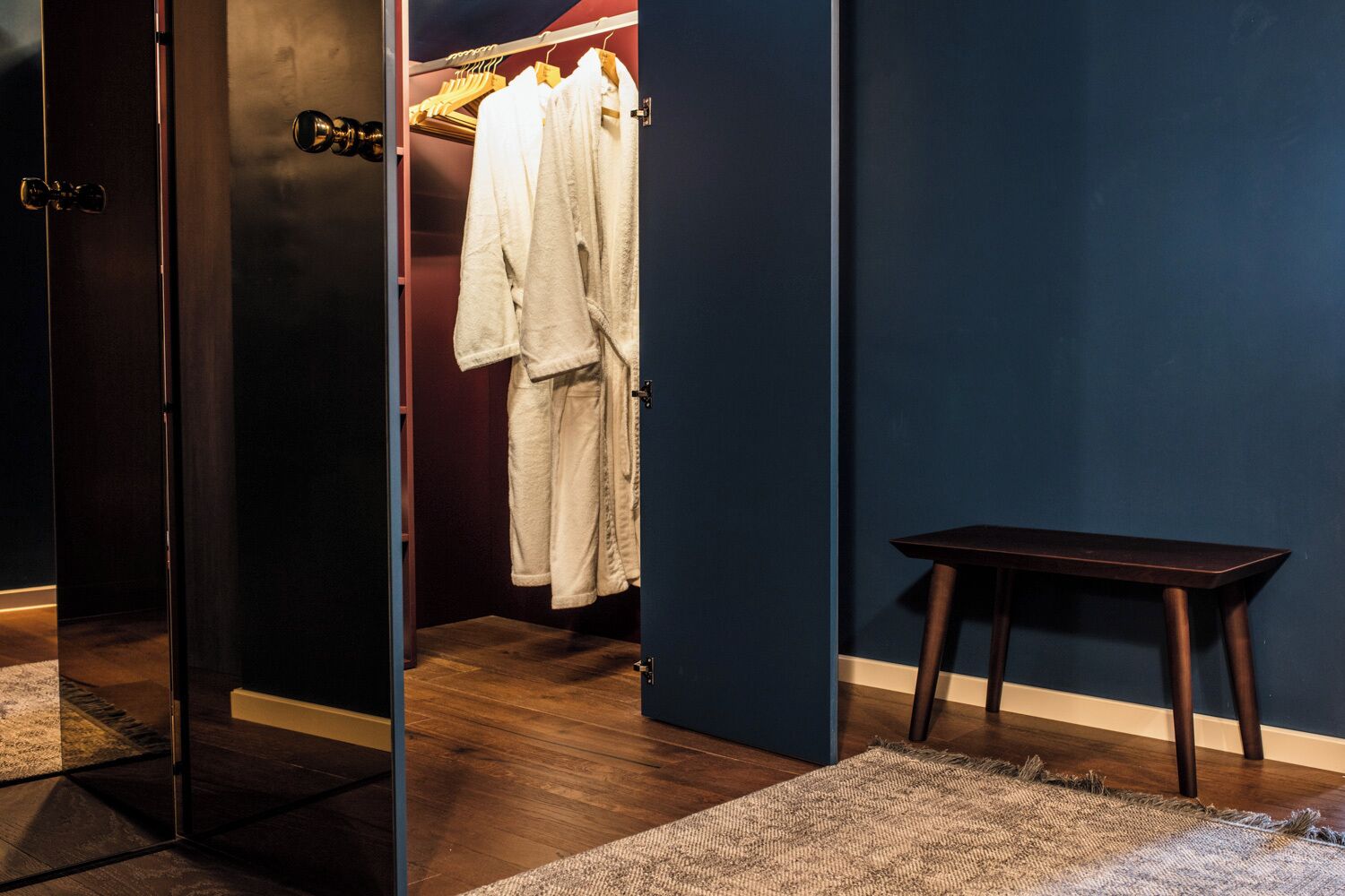A room with a wooden floor, a dark blue wall, a wooden bench, and a clothing rack with white robes.