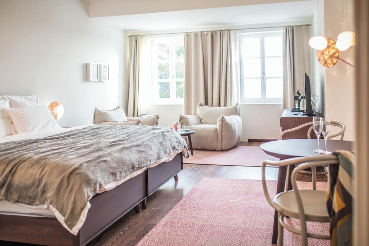 A hotel room with a bed, two chairs, a small table, a TV, and a window with curtains.