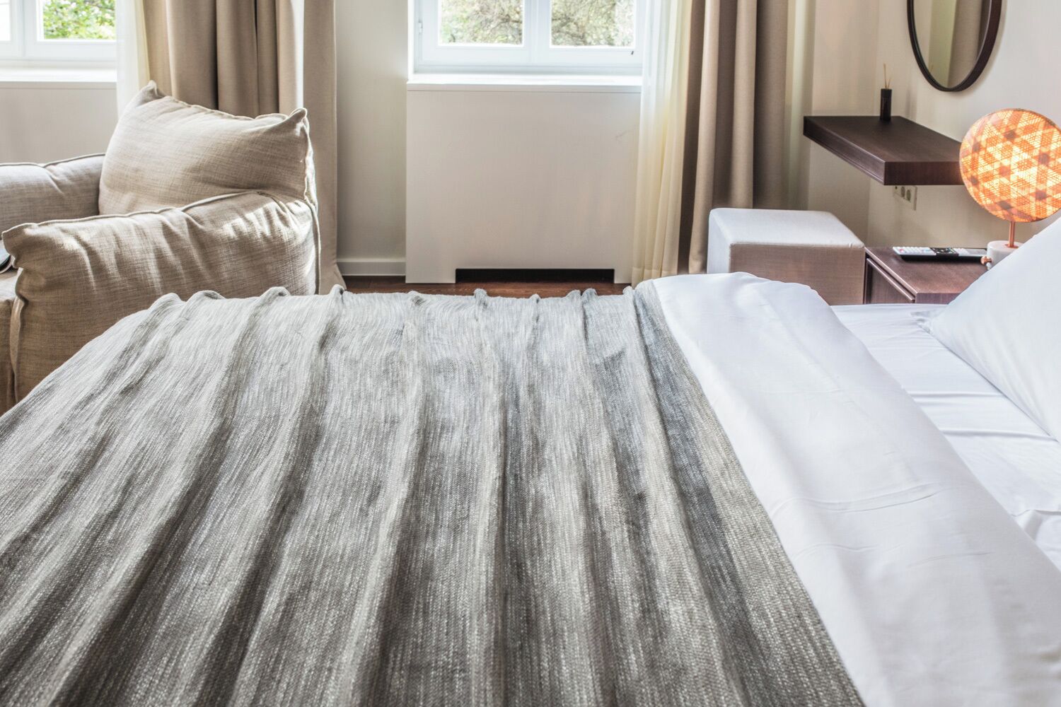 Quarto de hotel com cama, cadeira, mesa de cabeceira com abajur, espelho e janela com cortinas.