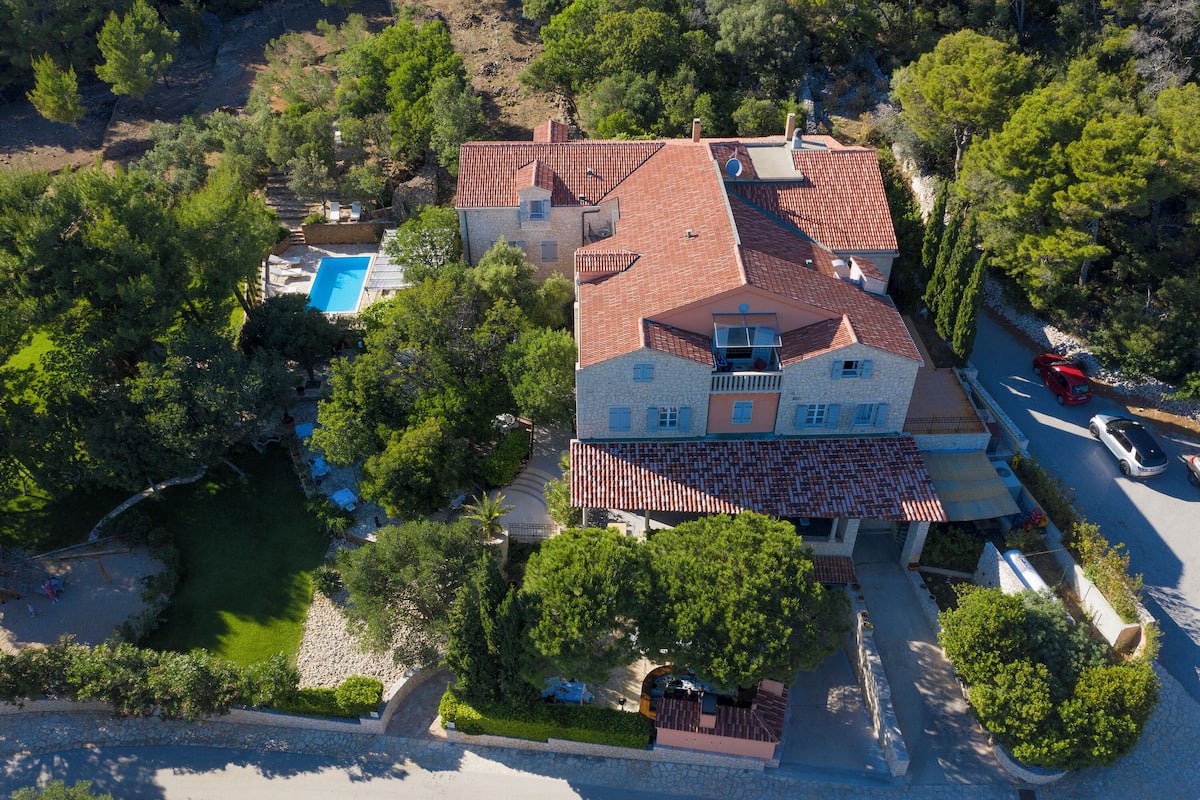 An aerial view of a villa with a swimming pool, surrounded by trees and a stone pathway.