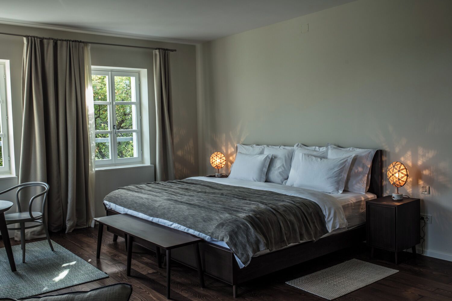 Un dormitorio con cama, mesitas de noche, una silla, un banco y una ventana.