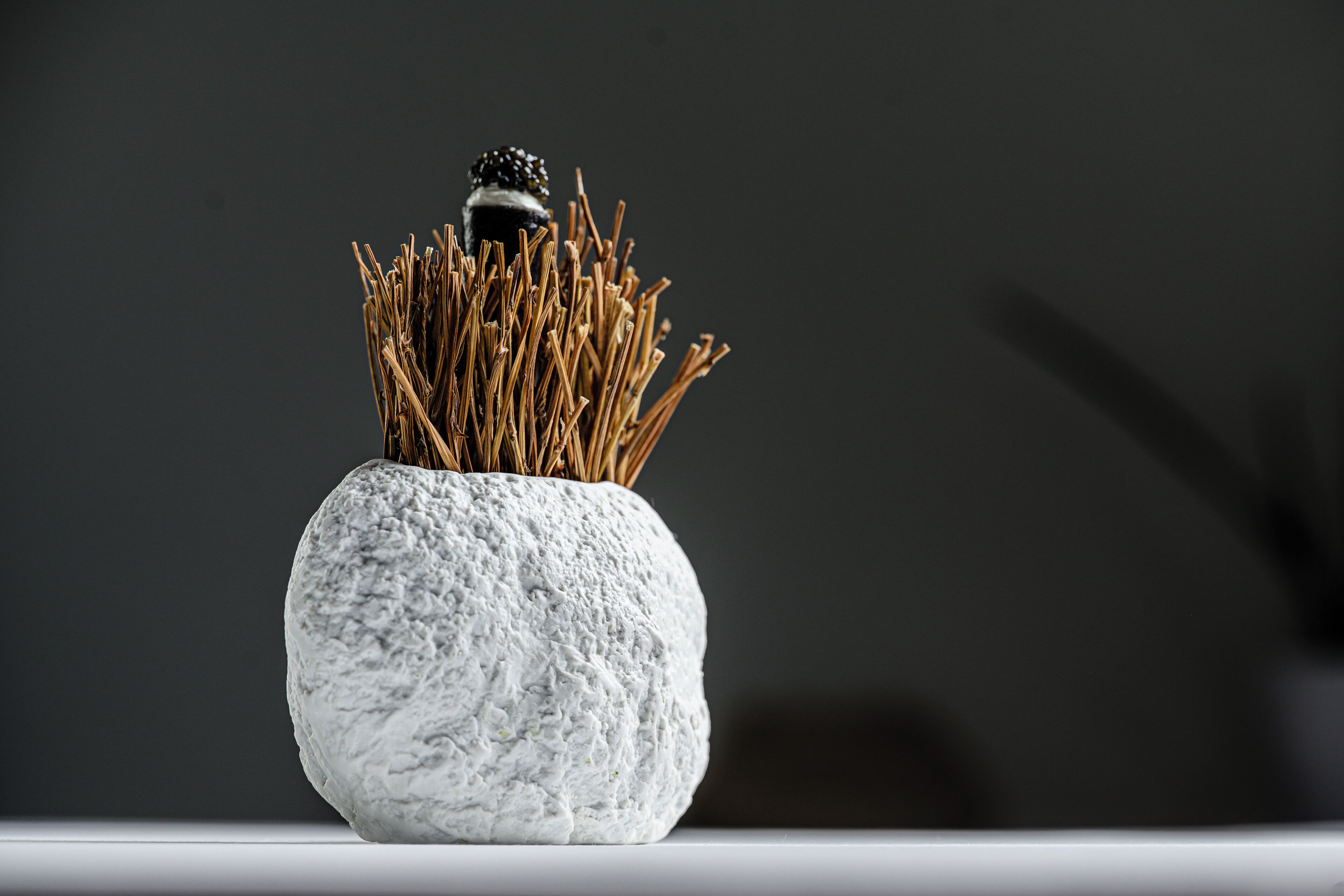 Un vase blanc à texture, surmonté d’un objet noir et rempli de roseaux séchés, le tout sur un fond sombre.