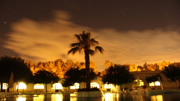 A nighttime view of a pool area with palm trees and lounge chairs, reflecting lights from surrounding buildings. - Laurito Resort & Spa (Oria)