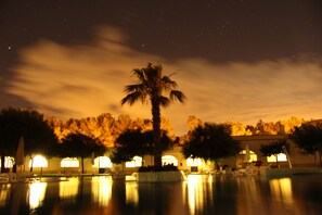 Vista nocturna de un área de piscina con palmeras y sillones reclinables, reflejando luces de edificios circundantes.
