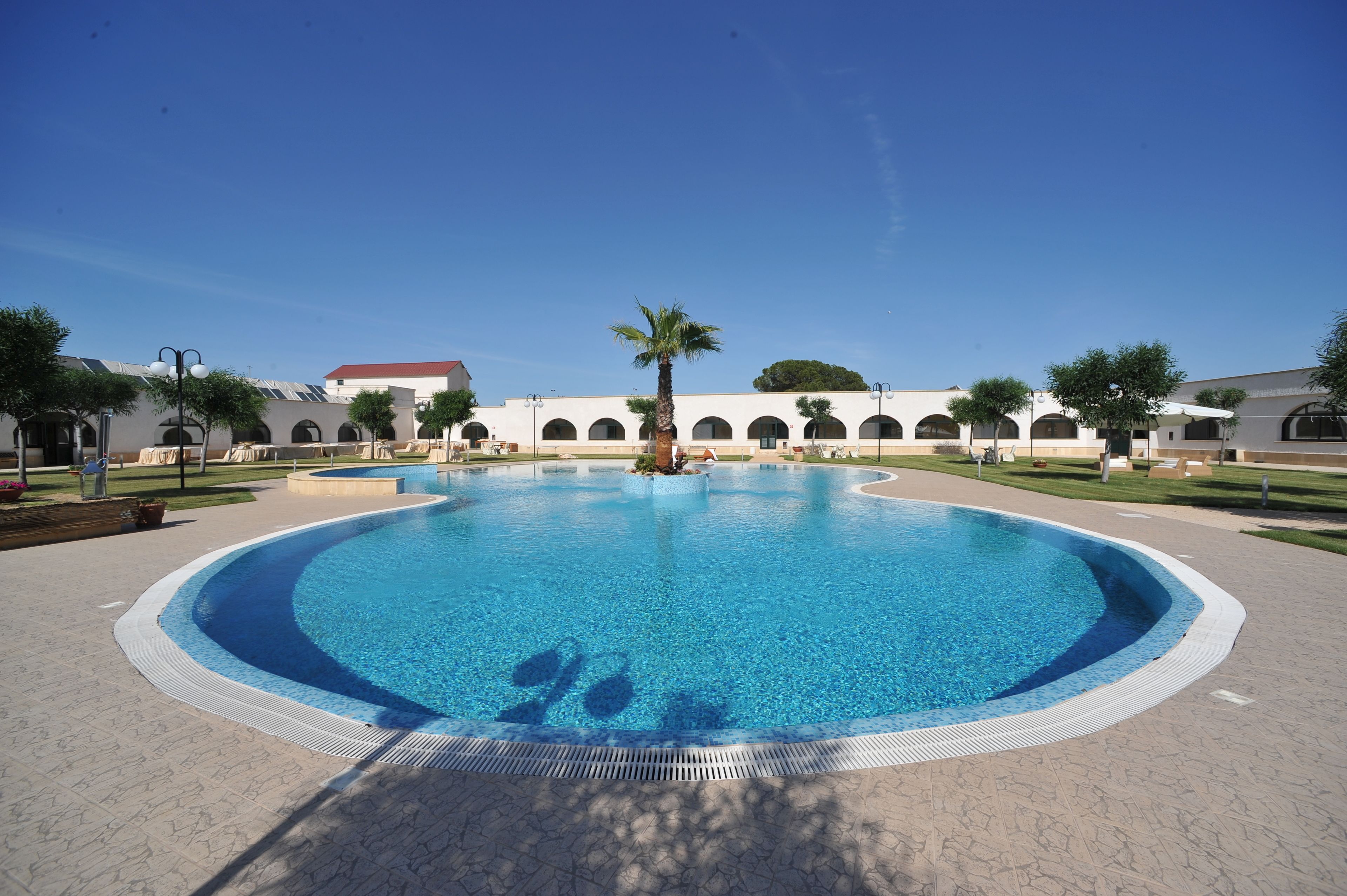 Una gran piscina circular con una palmera en el centro, rodeada por un edificio blanco con ventanas arqueadas y un techo rojo.