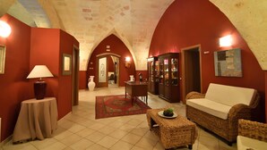 A hallway with a red wall, a tiled floor, a wicker sofa, a small table with a lamp, and a decorative rug. - Laurito Resort & Spa (Oria)