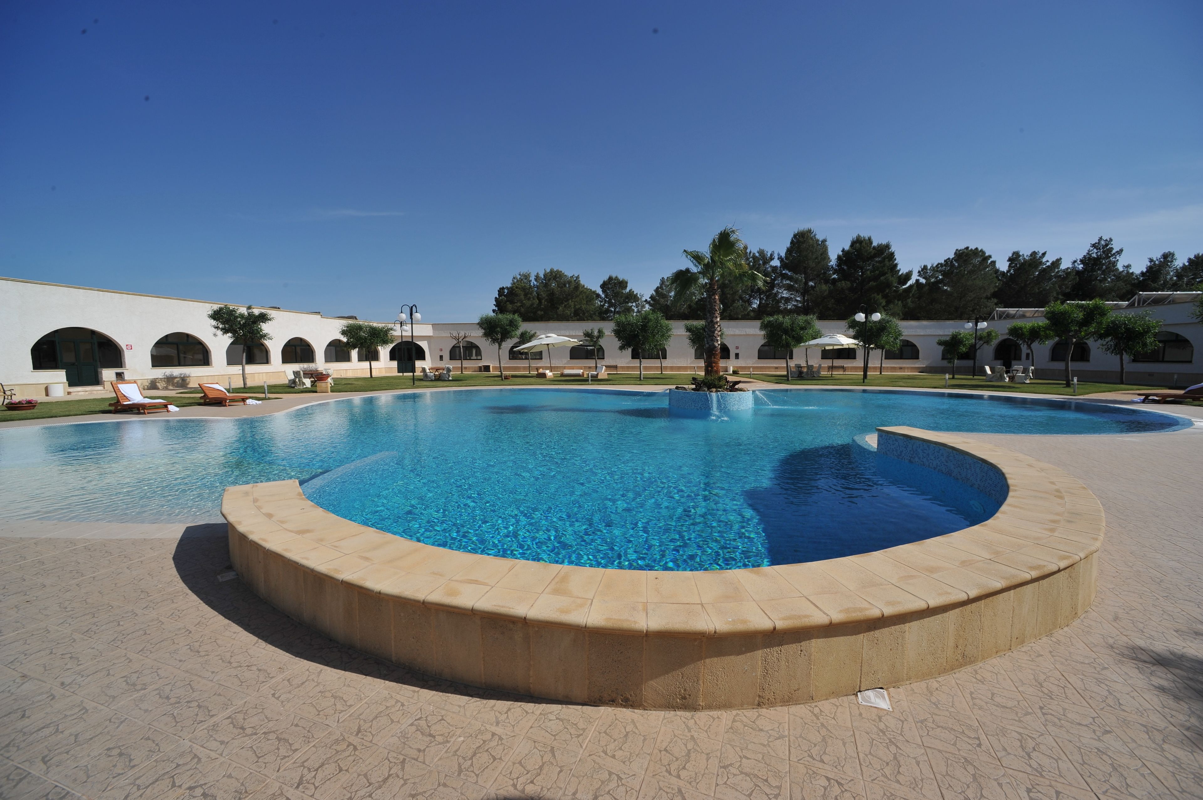 A large, clear swimming pool with a curved edge, surrounded by a paved walkway and palm trees.